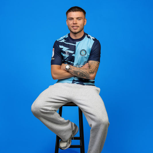 Man sitting on a stool wearing a blue patterned shirt with light grey pants against a blue background
